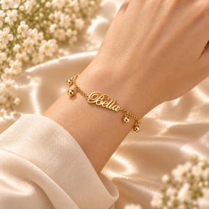 Può includere: Un bracciale a catena color oro con il nome Bella in carattere corsivo, arricchito da piccoli ciondoli sferici pendenti fissati a intervalli lungo le delicate maglie metalliche, creando un gioiello lucido e riflettente.