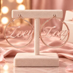 Può includere: Un paio di orecchini a cerchio in oro rosa con il nome Zoey in carattere corsivo integrato al centro di ogni cornice circolare. Gli orecchini sono esposti su un supporto beige a forma di T, mostrando una finitura metallica lucida.