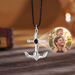 May include: A silver-toned anchor-shaped pendant featuring a central circular projection lens. The pendant hangs from a black chain. A circular light projection displays a photograph of a man carrying a young boy on his shoulders outdoors.