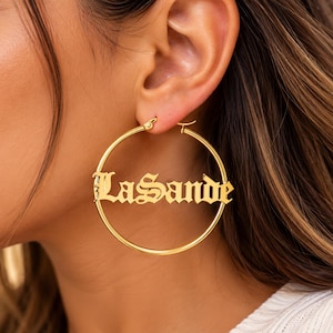 Può includere: Un orecchino a cerchio color oro con il nome LaSande scritto in un carattere gotico stilizzato al centro. Il cerchio è sottile e lucido, con la targhetta del nome integrata nel design, creando un accessorio audace.