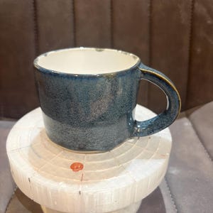 May include: A ceramic mug with a speckled dark blue exterior glaze and a smooth white interior. The mug features a sturdy, curved handle and a cylindrical body with a slightly flared rim, showcasing a handcrafted aesthetic with subtle colour variations.