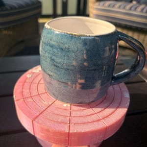 May include: A blue ceramic mug with a speckled, textured finish and a white interior, resting on a circular, segmented pink coaster with a shimmering surface. The mug features a rounded body and a sturdy handle, contrasting with the flat, pink base.