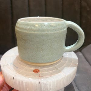 May include: A light sage green ceramic mug with a speckled texture and a smooth, rounded handle. The mug features a slightly flared rim and a plain, unglazed beige base. The surface has a matte finish with subtle indentations and variations.