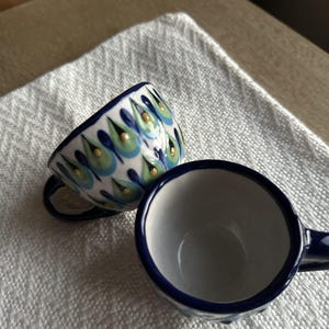 Pode incluir: Duas pequenas chávenas de café em cerâmica com base branca e um padrão pintado à mão de formas de gota azuis e verdes, acentuadas com pequenos pontos amarelos. Uma chávena está na vertical, enquanto a outra está inclinada de lado.
