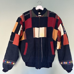 May include: A zip-up bomber jacket featuring a patchwork design of navy blue, maroon, and mustard yellow fabric panels. The sleeves include colorful woven trim details at the cuffs, and the jacket has two vertical side pockets and a ribbed collar.