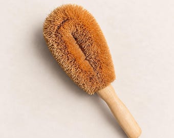 May include: A cleaning brush featuring a light-colored wooden handle and a large, oval-shaped head with stiff, brown natural fiber bristles arranged in a dense, textured pattern for scrubbing surfaces.