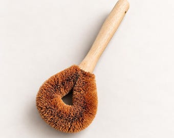 May include: A dish brush featuring a long, light-colored wooden handle and a teardrop-shaped head with stiff, brown, natural-looking bristles. The brush head has a central opening, and the handle includes a small hole at the end for hanging.