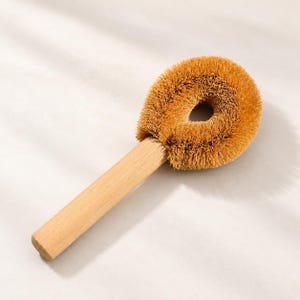 Puede incluir: Un cepillo para platos con mango de madera clara y cabezal circular con cerdas rígidas de color marrón de aspecto natural. El cabezal del cepillo tiene un centro hueco y las cerdas están dispuestas en una forma densa y redondeada.