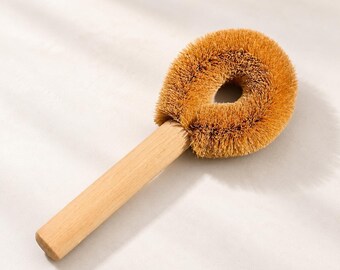 May include: A dish brush featuring a light-colored wooden handle and a circular head with stiff, brown, natural-looking bristles. The brush head has a hollow center, and the bristles are arranged in a dense, rounded formation for scrubbing.