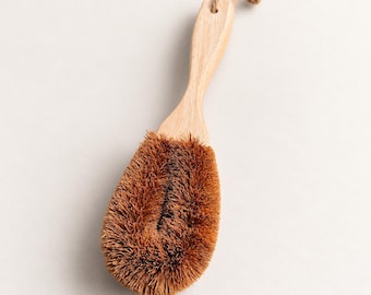 May include: A dish brush featuring a light-colored wooden handle and a thick, rounded head of stiff, brown natural fiber bristles. A thin, knotted jute twine loop is attached to the end of the handle for convenient hanging storage.