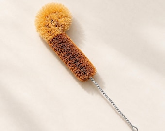 May include: A bottle brush featuring a long, twisted silver metal handle with a loop at the end. The head of the brush is composed of stiff, light brown and dark brown bristles arranged in a cylindrical shape with a rounded top.