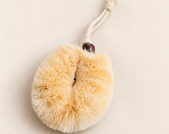 May include: A round, beige scrubbing brush with stiff, light-colored bristles arranged in a circular pattern. The brush features a dark brown wooden bead at the top, connected to a light-colored, braided cotton rope loop for hanging storage.