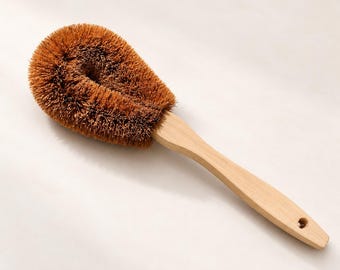 May include: A dish brush with a light-colored wooden handle and a round head featuring stiff, brown, natural-looking bristles. The handle is smooth and includes a small circular hole at the end for hanging, providing a functional tool for cleaning.