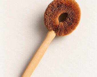 May include: A round dish brush with a light-colored wooden handle and stiff, brown, circular bristles arranged in a ring shape. The brush features a central opening within the dense, textured bristle head, attached to a smooth, cylindrical handle.