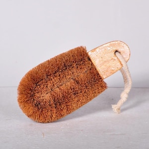 Puede incluir: Un cepillo de fregar rectangular con cerdas naturales rígidas de color marrón, fijadas a un mango de madera clara. Un lazo grueso de cuerda de algodón de color blanco roto atraviesa un orificio en el mango para colgarlo.