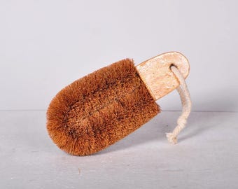 May include: A rectangular scrubbing brush featuring stiff, brown natural fiber bristles attached to a light-colored wooden handle. A thick, off-white cotton rope loop is threaded through a hole in the handle for hanging purposes.