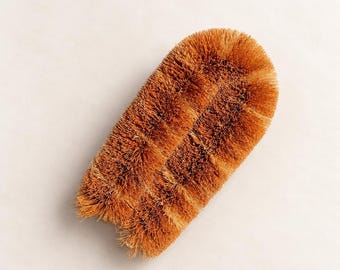 May include: A U-shaped scrubbing brush featuring dense, stiff, light brown natural fibers. The brush has a rounded top and two parallel bristle sections that meet at the base, creating a compact, functional tool for cleaning tasks.