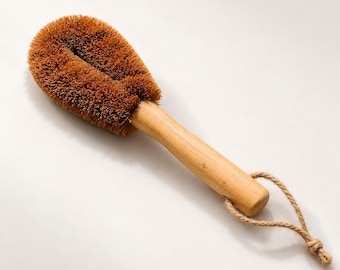 May include: A cleaning brush with a light-colored wooden handle and a rounded head of stiff, brown, natural-looking bristles. A short, braided jute rope loop is attached to the end of the handle for hanging, providing a simple and functional design.
