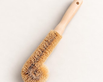 May include: A dish brush with a long, light-colored wooden handle featuring a small circular hole at the end. The brush head consists of stiff, tan-colored natural bristles arranged in a cylindrical shape that curves into a rounded end.
