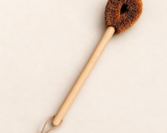 May include: A long-handled cleaning brush featuring a light-colored wooden handle and a round, brown, stiff-bristled head. A thin, knotted twine loop is attached to the base of the handle for hanging, providing a simple and functional design.