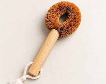 May include: A round dish brush with stiff brown bristles and a light-colored wooden handle. The handle features a white braided cotton loop at the base with a small tassel, providing a functional grip for hanging the cleaning tool.