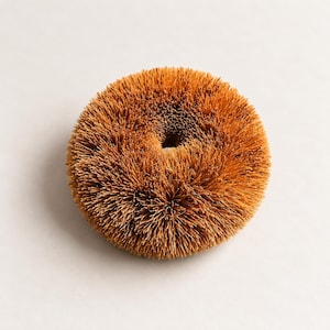 Puede incluir: Un cepillo de fregar redondo de color tostado con cerdas densas y rígidas dispuestas en un patrón circular con un pequeño centro hueco. La textura es gruesa y fibrosa, creando una apariencia esférica uniforme con una superficie plana.