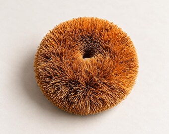 May include: A round, tan-colored scrubbing brush featuring dense, stiff bristles arranged in a circular pattern with a small, hollow center. The texture is coarse and fibrous, creating a uniform, spherical appearance with a flat top surface.