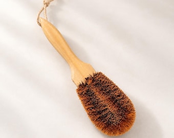 May include: A cleaning brush with a light-colored wooden handle and a thick, rounded head of stiff, dark brown bristles. A short, twisted fiber loop is attached to the end of the handle for hanging, providing a simple and functional design.