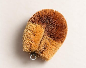 May include: A round scrubbing brush made of stiff, light and dark brown natural fibers. The brush features a dense, circular arrangement of bristles and includes a small, circular metal loop at the base for hanging or storage purposes.