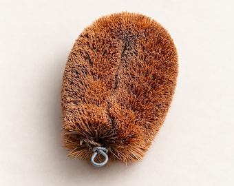 May include: An oval-shaped scrubbing brush made of stiff, brown natural coconut coir fibers. The brush features a dense, textured surface and includes a small, circular silver-colored metal loop at the bottom edge for hanging or storage purposes.