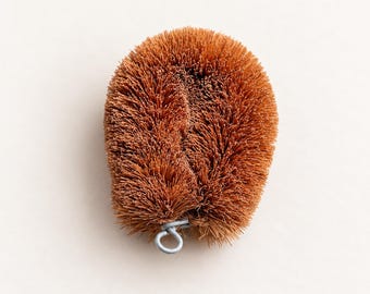 May include: A round, brown scrubbing brush made of stiff, fibrous bristles. The brush features a dense, textured surface and includes a small, silver-colored metal loop at the bottom edge for hanging or storage purposes.