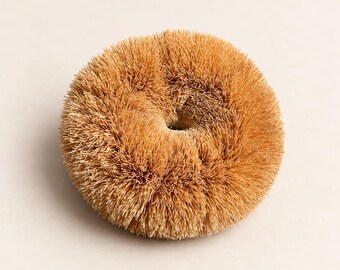 May include: A round, tan-colored scrubbing brush with a dense, circular arrangement of stiff, natural-looking bristles. The brush features a hollow center, creating a donut-like shape with a textured, fibrous surface throughout the entire object.