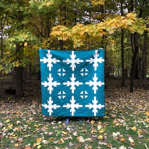 May include: A teal blue quilt with a white snowflake pattern. The quilt is folded and displayed in front of a background of fall foliage.