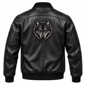 May include: A black leather bomber jacket featuring a ribbed collar, cuffs, and hem. The back of the jacket displays a large, detailed graphic patch of a wolf head with piercing red eyes, rendered in shades of grey, black, and white.