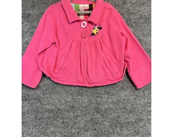 Puede incluir: Una chaqueta de color rosa brillante con mangas largas, cuello ancho, dos botones frontales y dos bolsillos laterales. La prenda incluye un bordado del personaje Minnie Mouse en el pecho derecho y un forro de rayas coloridas en el cuello.