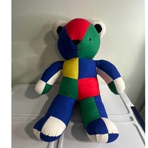 Può includere: Un orsacchiotto di peluche realizzato con toppe di tessuto multicolore in rosso, verde, blu, giallo e bianco. L'orso presenta una testa rotonda con occhi e naso cuciti in nero, orecchie arrotondate e un corpo segmentato con arti in stile patchwork.