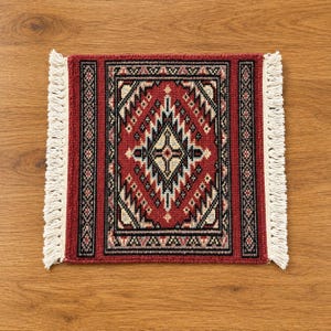 May include: A rectangular woven rug featuring a deep red background with a central diamond pattern in black, white, and light blue. The rug is framed by geometric borders and finished with off-white fringe along the two shorter side edges.