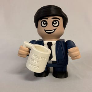 May include: A plastic figurine of a person with dark hair, wearing a navy blue suit with a white shirt and black tie. The figurine holds a white mug in its right hand with the text "WORLD'S BEST BOSS" embossed on the front in capital letters.