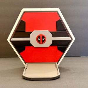 May include: A hexagonal display stand featuring a vibrant red background with black geometric accents. A central light gray rectangular plate displays a circular red and black Deadpool character logo. The base is a light gray platform.