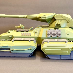 May include: A light green and gray plastic model tank featuring a long main cannon, a turret with angular paneling, and two separate track assemblies. The vehicle includes a small mounted weapon on the upper hull and rectangular side armor plates.