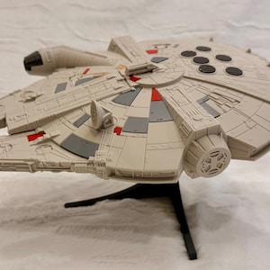 May include: A plastic model of the Millennium Falcon starship featuring a light gray hull with dark gray and red accent panels. The circular spacecraft has a cockpit on the side, a top-mounted radar dish, and six black circular vents on the rear.