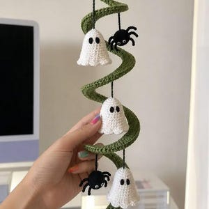 May include: A spiral-shaped green crochet hanging decoration featuring three white crocheted ghosts and two black crocheted spiders suspended from the structure by thin black strings. Each ghost has two black eyes and each spider has white googly eyes.