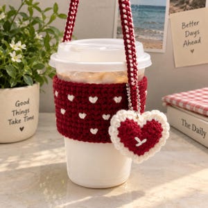 May include: A clear plastic iced coffee cup featuring a dark red crochet sleeve with a repeating white heart pattern. The cup includes a matching dark red and white crochet carrying strap and a dangling heart-shaped crochet charm with a white border.