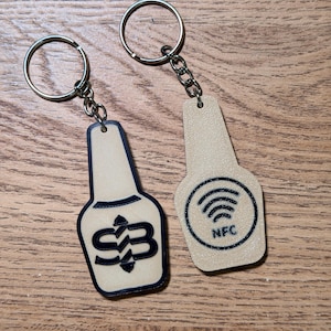 Pode incluir: Dois porta-chaves bege em forma de garrafa com argolas de metal. Um apresenta um logótipo preto com as letras SB e setas, enquanto o outro exibe um ícone circular preto com linhas curvas e o texto NFC, indicando conectividade sem fios.
