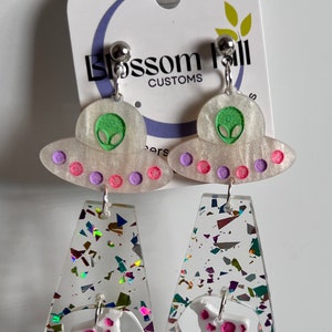 May include: A pair of dangle earrings featuring a white UFO charm with a green alien face and pink and purple accents. The bottom charm is a clear, iridescent, glittery rectangle with a white cow charm with pink spots.