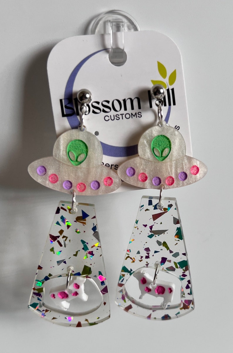 May include: A pair of dangle earrings featuring a white UFO charm with a green alien face and pink and purple accents. The bottom charm is a clear, iridescent, glittery rectangle with a white cow charm with pink spots.