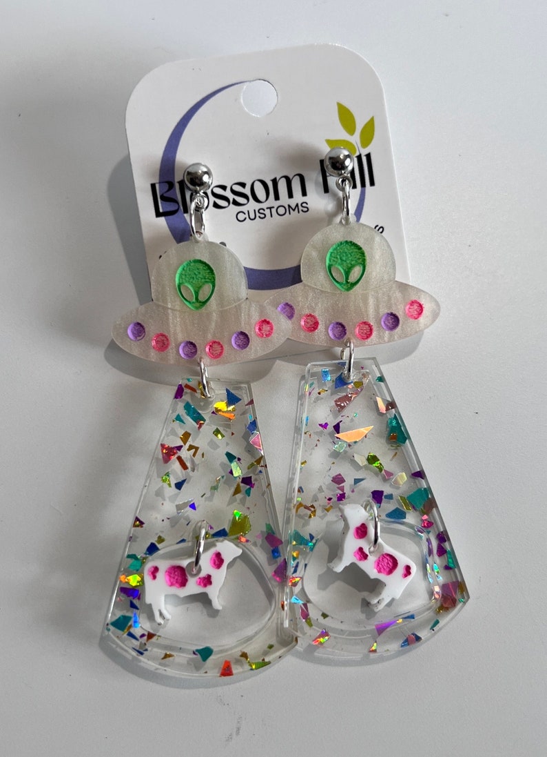 May include: A pair of dangle earrings featuring a white UFO with a green alien face and pink and purple dots. The UFO is attached to a clear, white, and iridescent glitter-filled dangle with a white cow silhouette with pink spots.