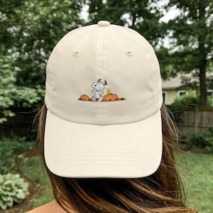 May include: A beige baseball cap featuring an embroidered graphic of the cartoon character Snoopy sitting between two orange pumpkins with a small yellow bird, Woodstock, standing on one of the pumpkins, all surrounded by scattered autumn leaves.
