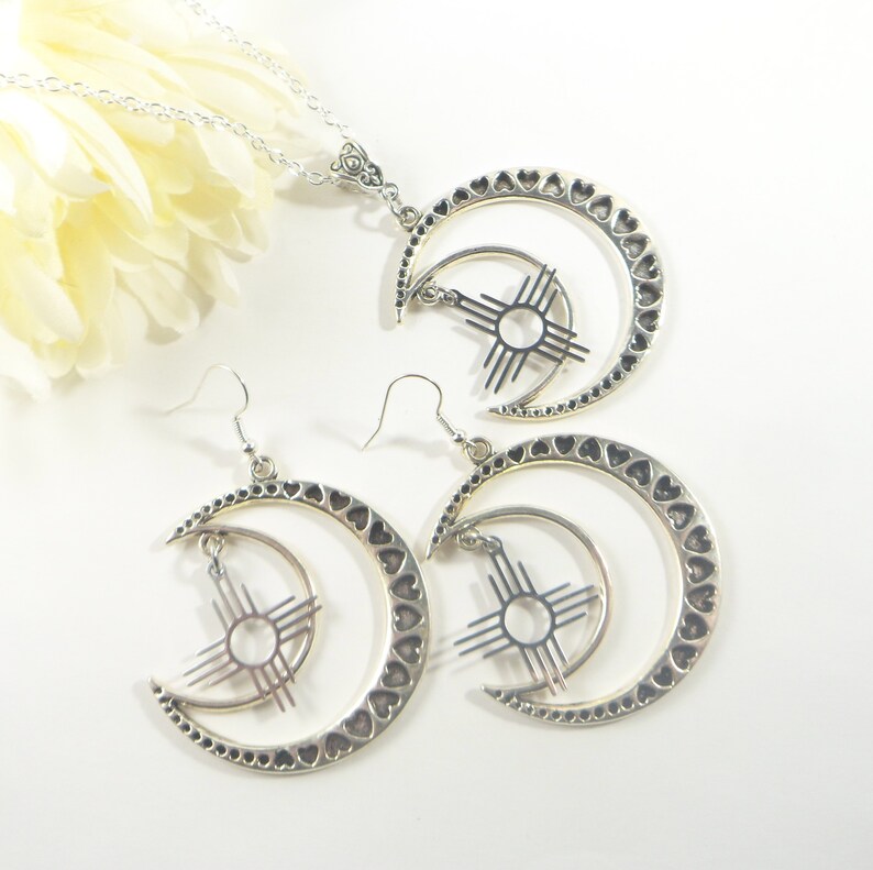 May include: A silver-toned jewelry set featuring a necklace and a pair of earrings. Each piece showcases a crescent moon design with heart-shaped cutouts and a sun symbol. The necklace has a delicate chain.