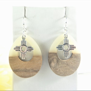 Puede incluir: Un par de pendientes de madera con un dije de plata en forma del s&iacute;mbolo Zia. Los pendientes son de forma ovalada y tienen un patr&oacute;n de veta de madera marr&oacute;n claro y blanco.